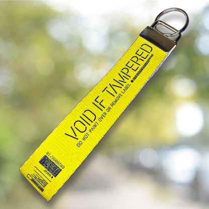 May include: A yellow security strap with a metal ring. The strap has black text that reads 'VOID IF TAMPERED' and 'DO NOT PAINT OVER OR REMOVE LABEL'. A barcode and other text are printed on the strap. The strap is angled against a background with a blur decorations.