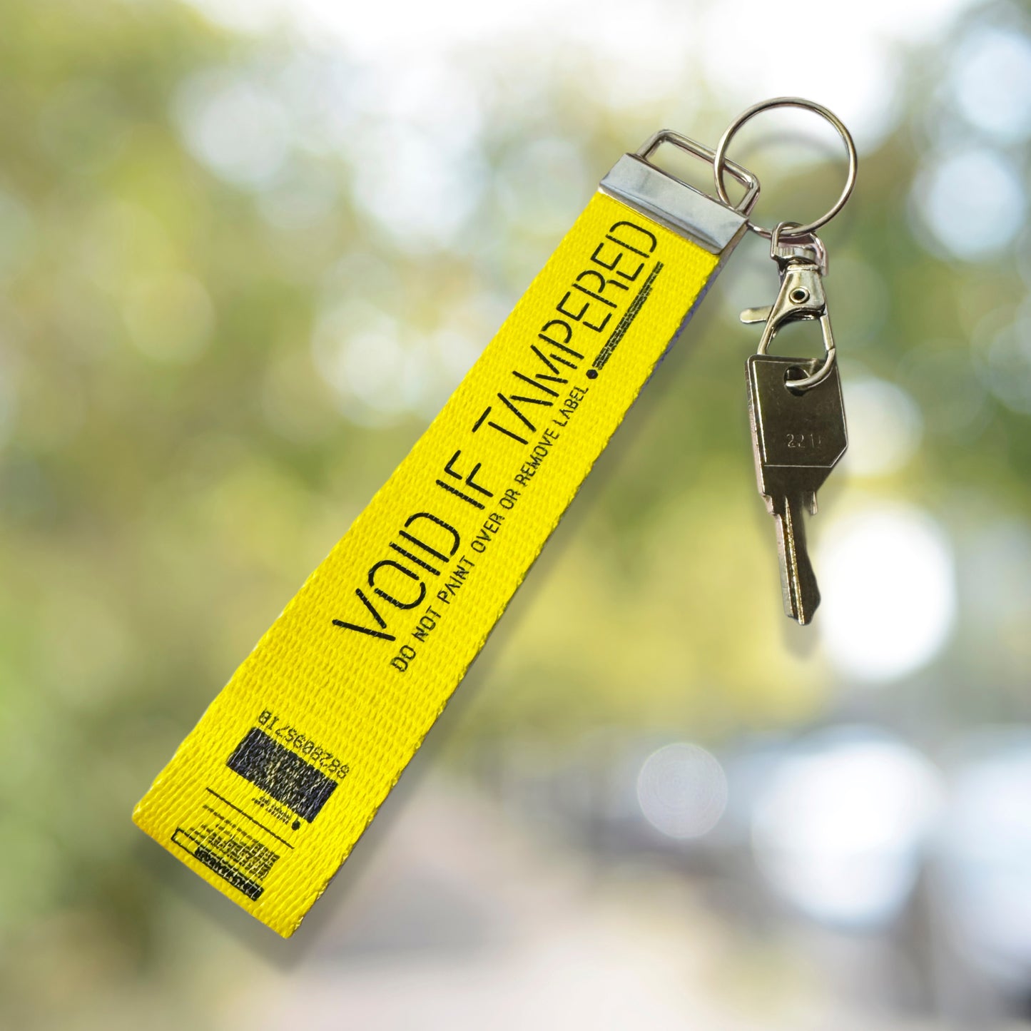 May include: A yellow lanyard with the words 'VOID IF TAMPERED' printed in black. The lanyard has a metal clasp and key ring attached, with a key hanging from the ring. The lanyard also has a black and white barcode and text printed on it. 